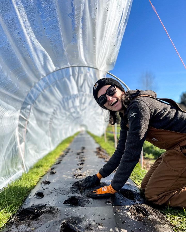A woman kneeling over a raised bed with overalls and gardening gloves while smiling into the camera. 