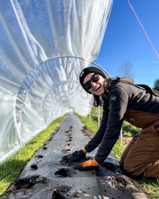 A woman kneeling over a raised bed with overalls and gardening gloves while smiling into the camera. 